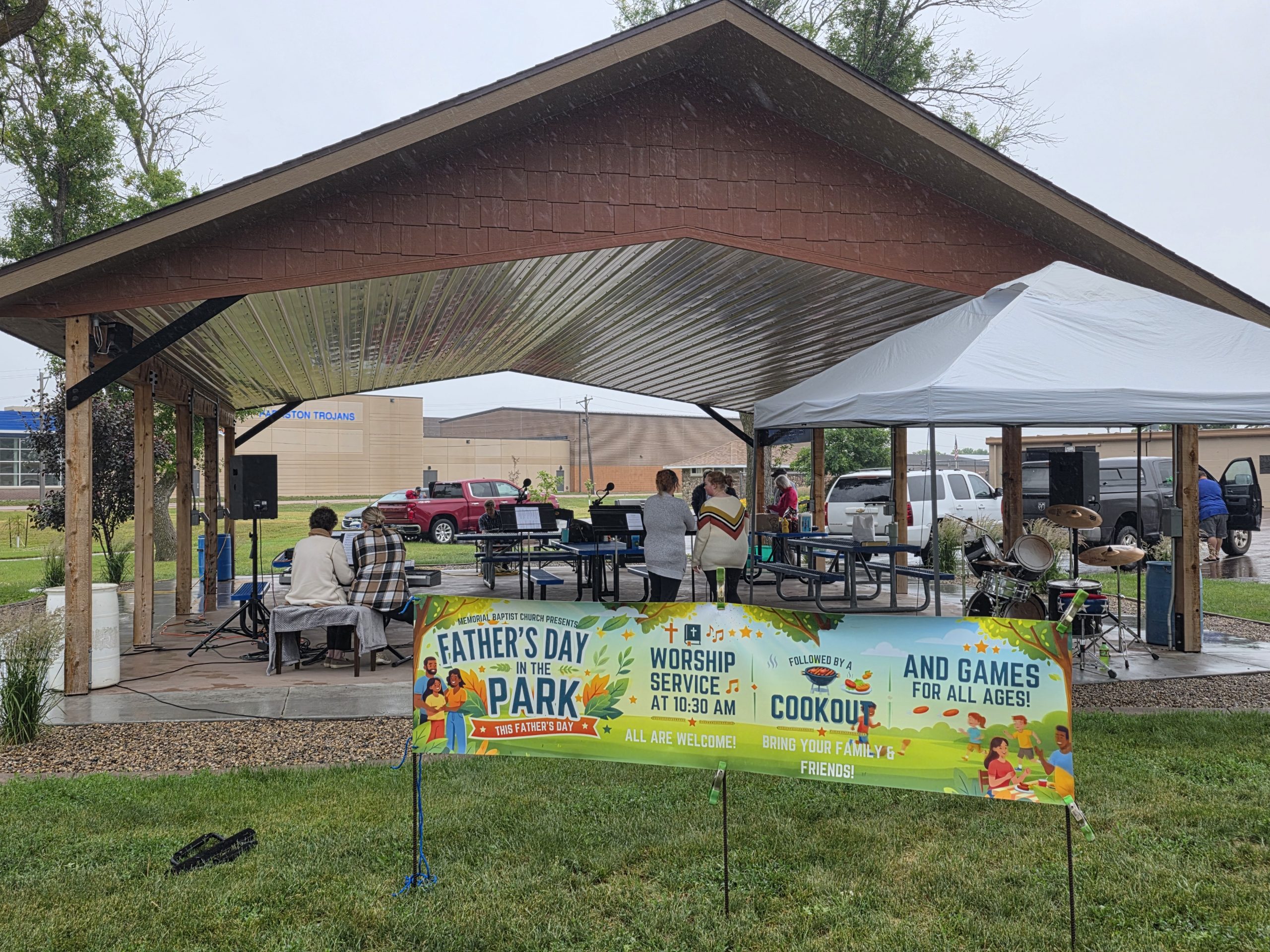 A banner reads "Father's Day in the Park" beside a stage and drum kit under a pavilion