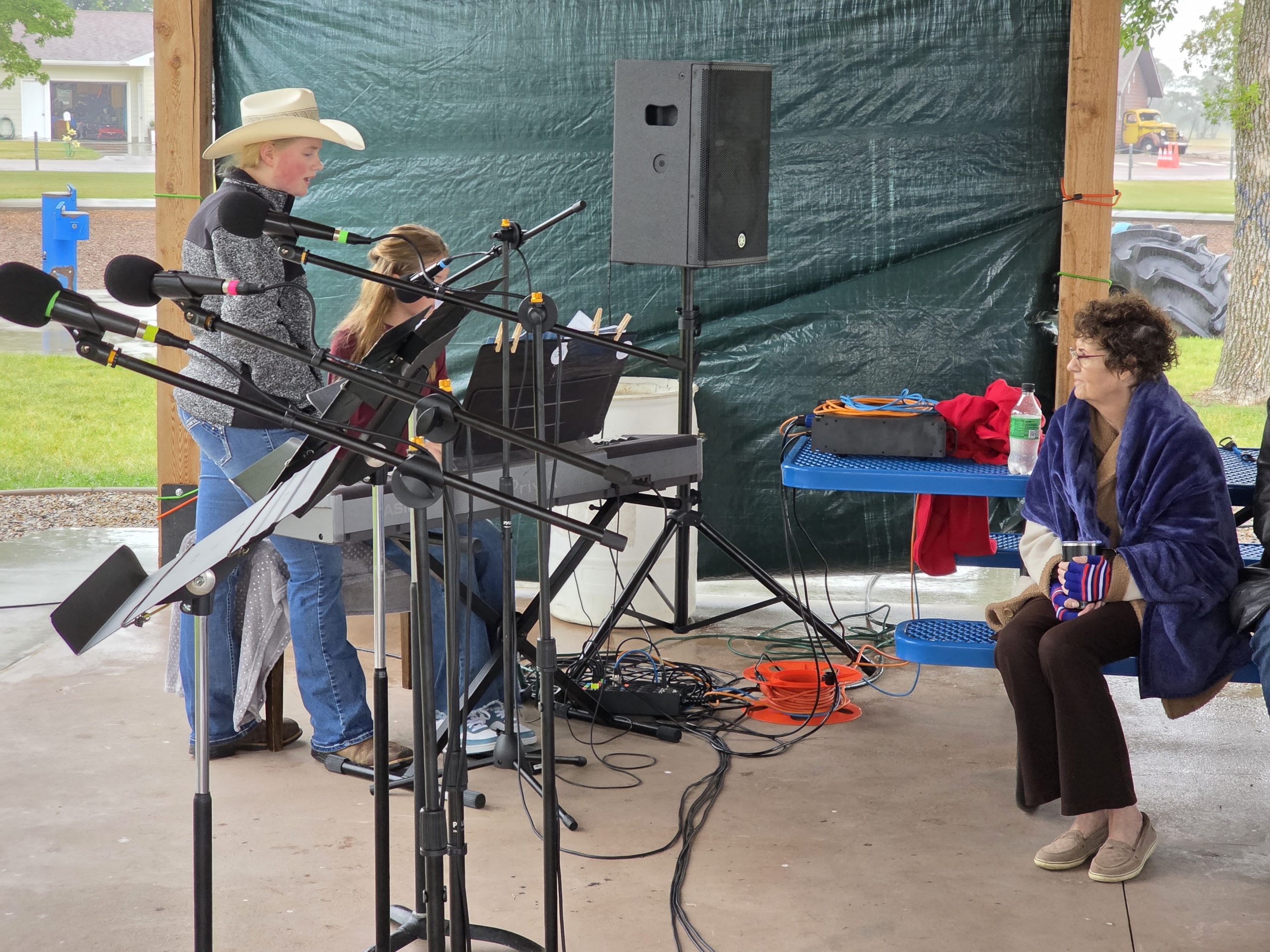 Sarah Boyle sings in a cowboy hat while Olivia Boyle plays piano under the pavilion
