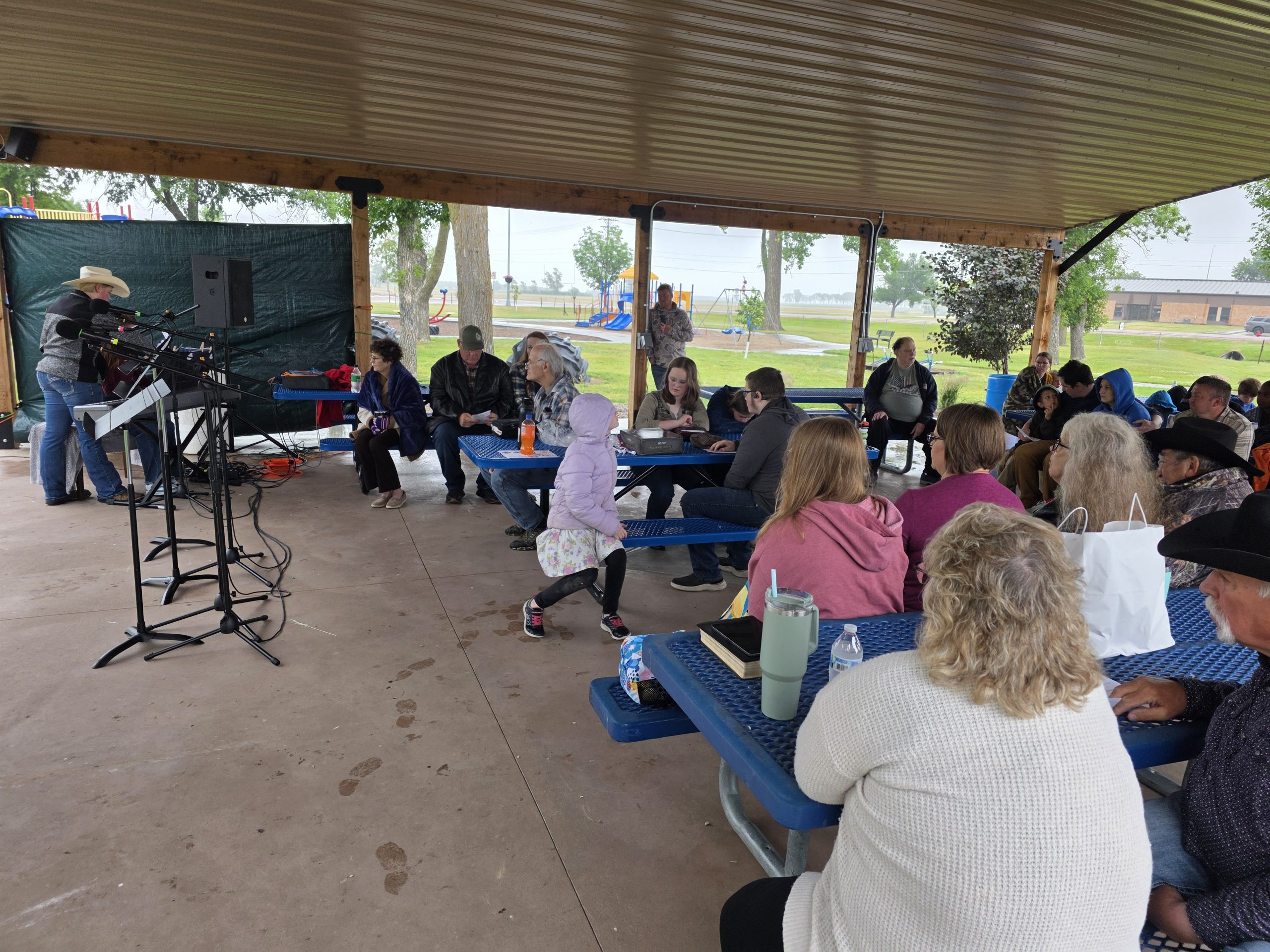 About fifty people sit at blue picnic tables with Bibles, listening under the pavilion