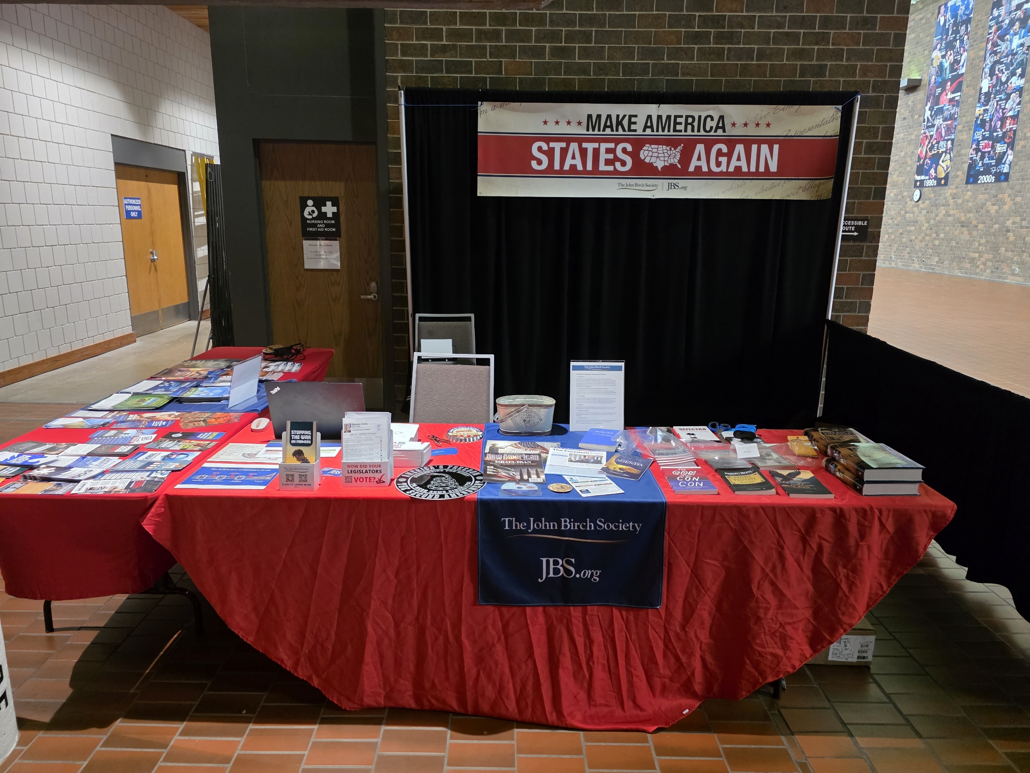 The John Birch Society informational booth at the 2026 R-CALF USA convention in Rapid City, with literature spread across a red table and a 'Make America States Again' banner.
